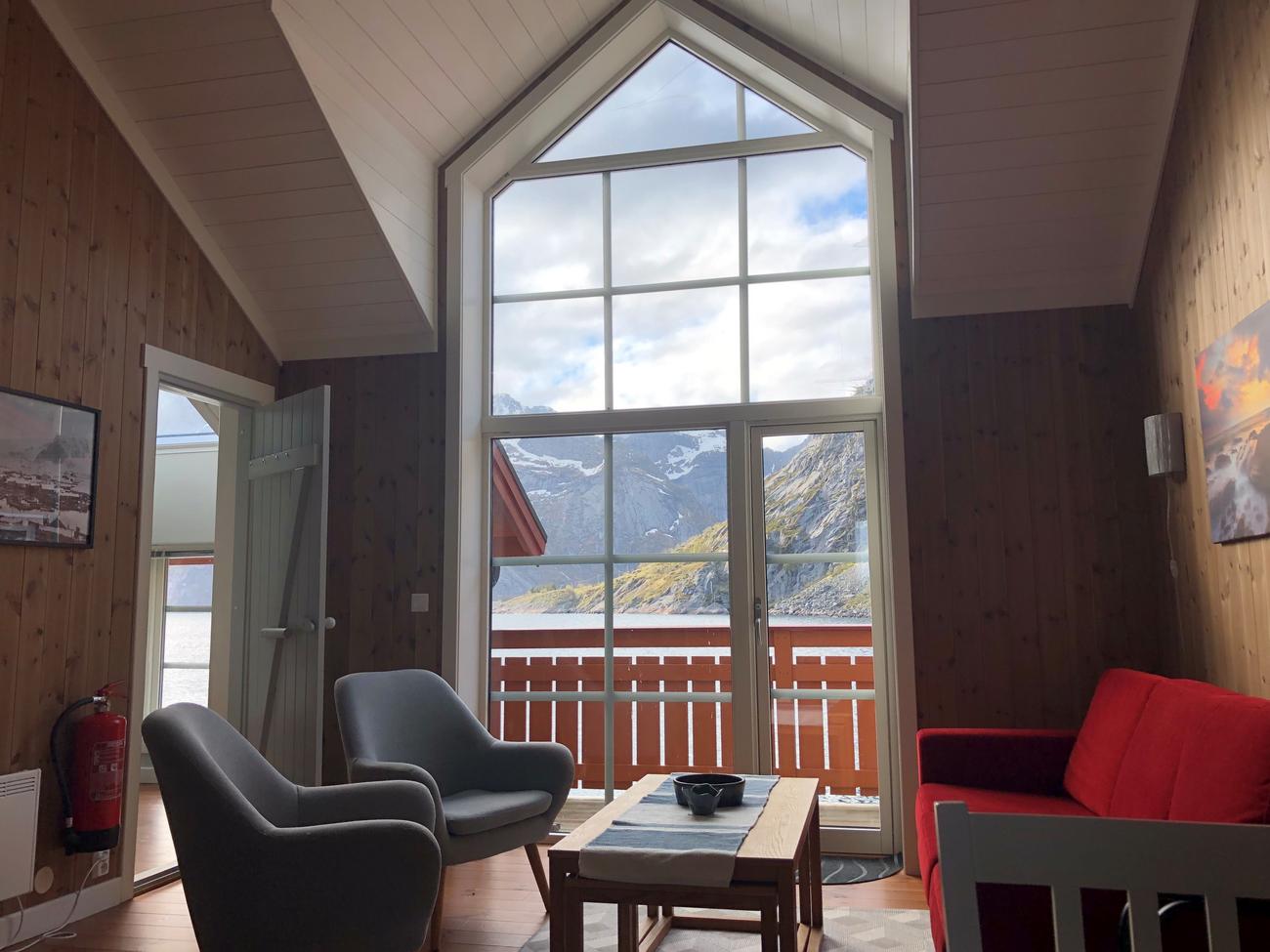 Stue med karakteristisk gulv-til-tak vindu ut mot Reinefjorden. Salong med sofa, bord og to stoler hvor man kan nyte Lofotens beste utsikt. 