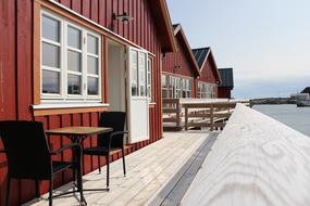 Fire r&oslash;de rorbuen med terrasse over vannet. Et bord og to stoler p&aring; en terrasse.