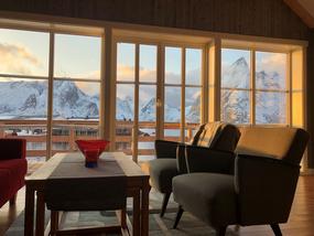 Stue med r&oslash;d sofa, gode stoler og sofabord. Hel vindusfasade i bakgrunn med balkongd&oslash;r og terrasse, med utsikt mot Reinefjorden og hvitkledd fjellrekke.
