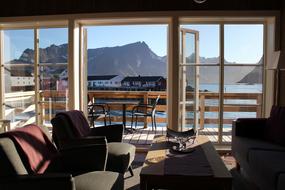 Stue med utsikt over Hamn&oslash;y havn og Reinefjorden. En sofa, et bord og to lenestoler foran de &aring;pne d&oslash;rene til terrassen.