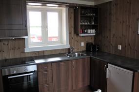 Kj&oslash;kken med komfyr, kj&oslash;leskap og oppvaskmaskin. Hyller over for oppbevaring av kopper og glass. Te-koker, presskanne og br&oslash;drister p&aring; benken. Skuffer og skap under som er fult utstyrt med kj&oslash;kkenredskaper. Vindu over vask og koketopp ved siden av vindu