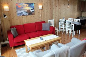 Stue med sofa, bord og lenestoler. Spisebord med plass til seks personer. Lampetter p&aring; vegg og bilde av strand i Lofoten over sofa. Leselys i hj&oslash;rnet 